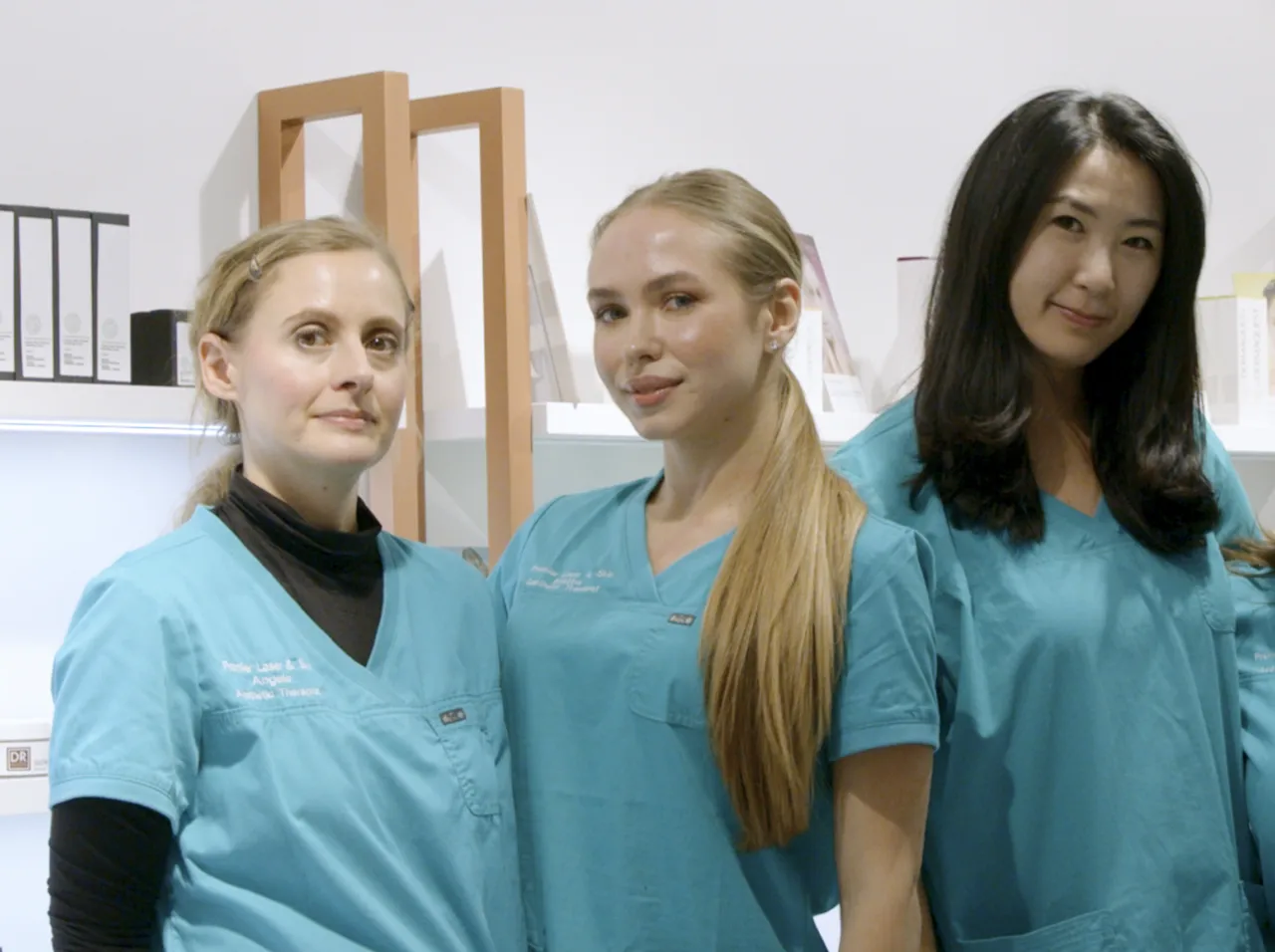 Three women stand side-by-side in light blue V-neck uniform tops featuring embroidered text that includes the words "Premier Laser & Skin.